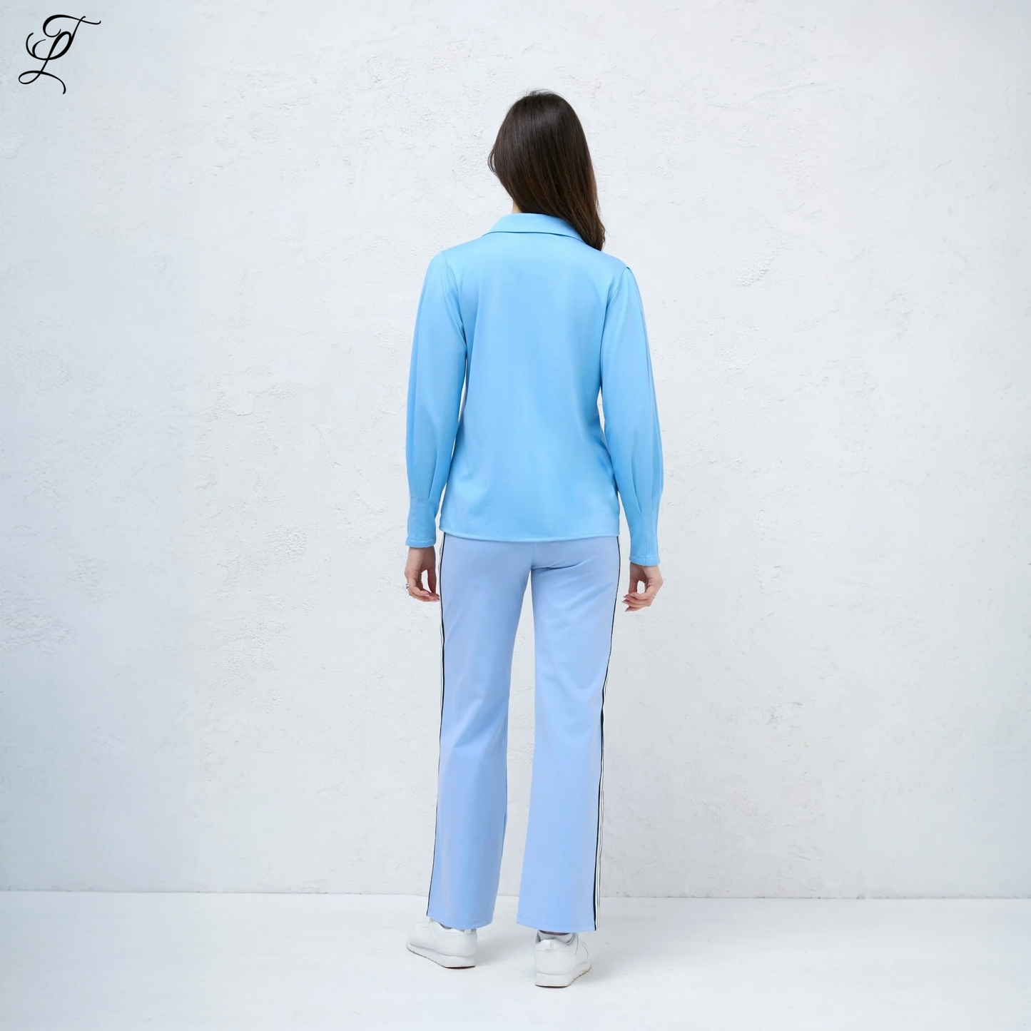 Chastar Stretch Blouse in lichtblauw, getoond vanaf de achterkant met elegante pofmouwen. Model draagt de blouse gecombineerd met een matching broek en witte sneakers tegen een witte achtergrond. Perfect voor een stijlvolle casual look. Blouses, Collectie, Bestsellers.