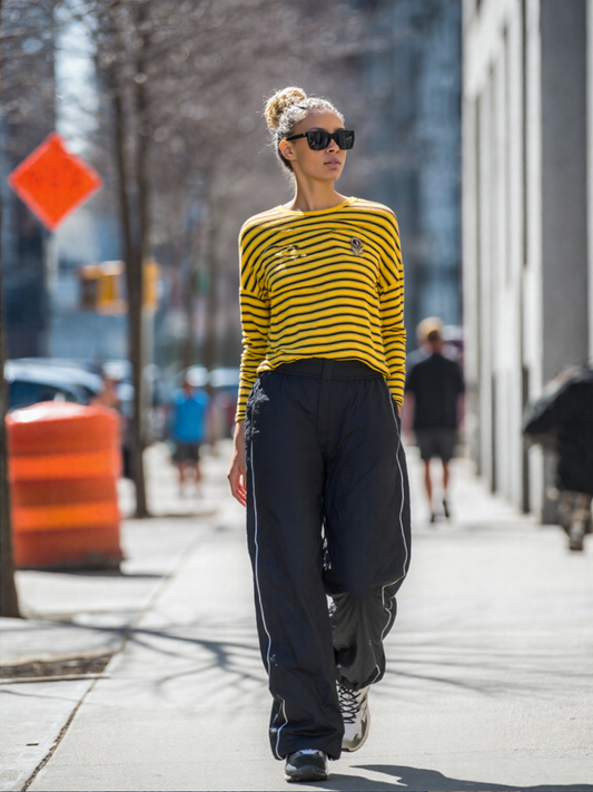 Vrouw loopt op straat met gele gestreepte lange mouw top en zwarte wijde broek in stedelijke omgeving.